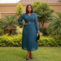 Nigerian female model wearing teal pleated midi gown with sheer sleeves and crystal belt in upscale garden setting