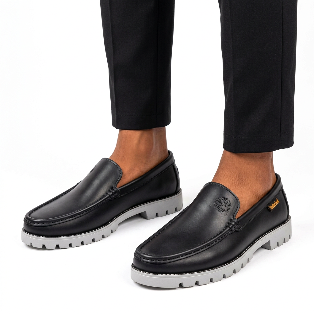 Male model wearing black embossed-emblem grey lug-sole loafers, both shoes visible on white