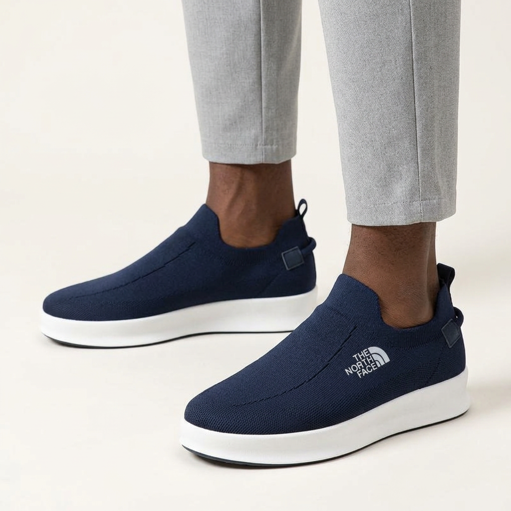 Male model wearing navy knit slip-ons with grey trousers, both shoes visible on an off-white background