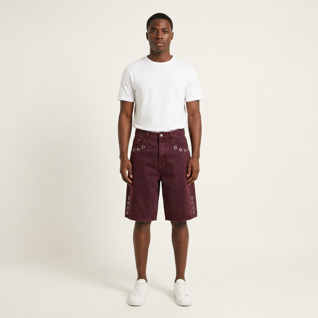 Male model wearing burgundy eyelet denim shorts with a white tee and trainers on a plain off-white background