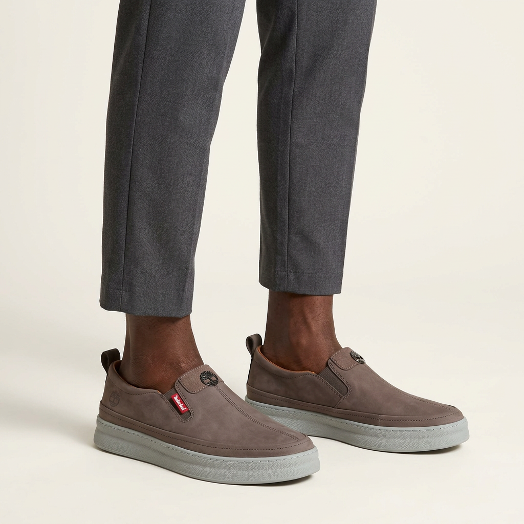 Both feet wearing grey-taupe slip-on shoes against a plain off-white background