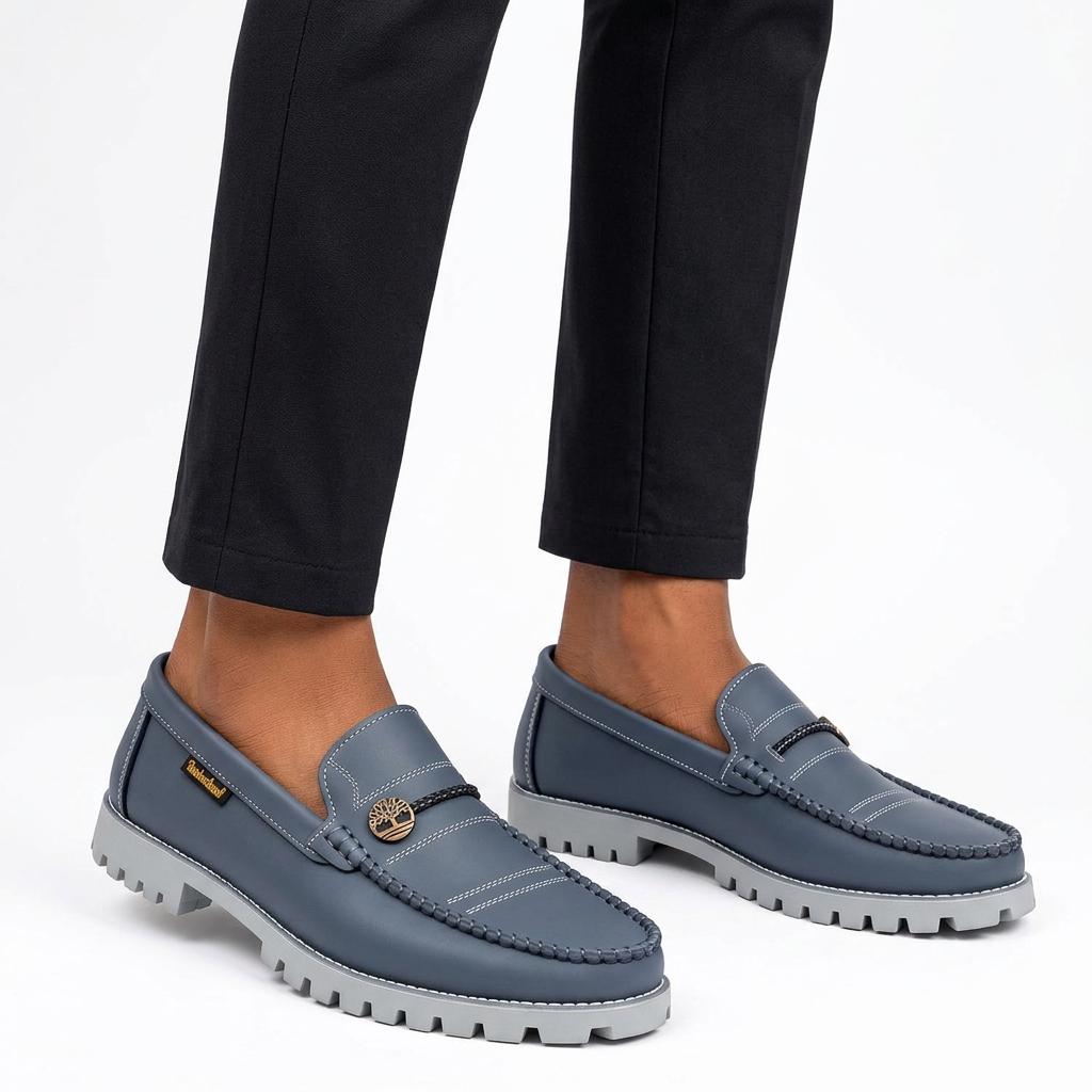 Male model wearing slate-blue chain-emblem loafers with both shoes visible on white
