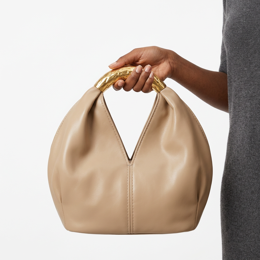 Warm-cream rounded handbag without label held by one hand at its gold-tone handle