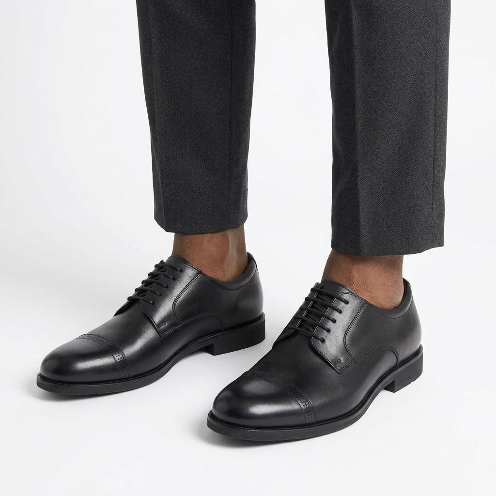 Plain black leather cap-toe derby shoes worn by a male model, both feet visible