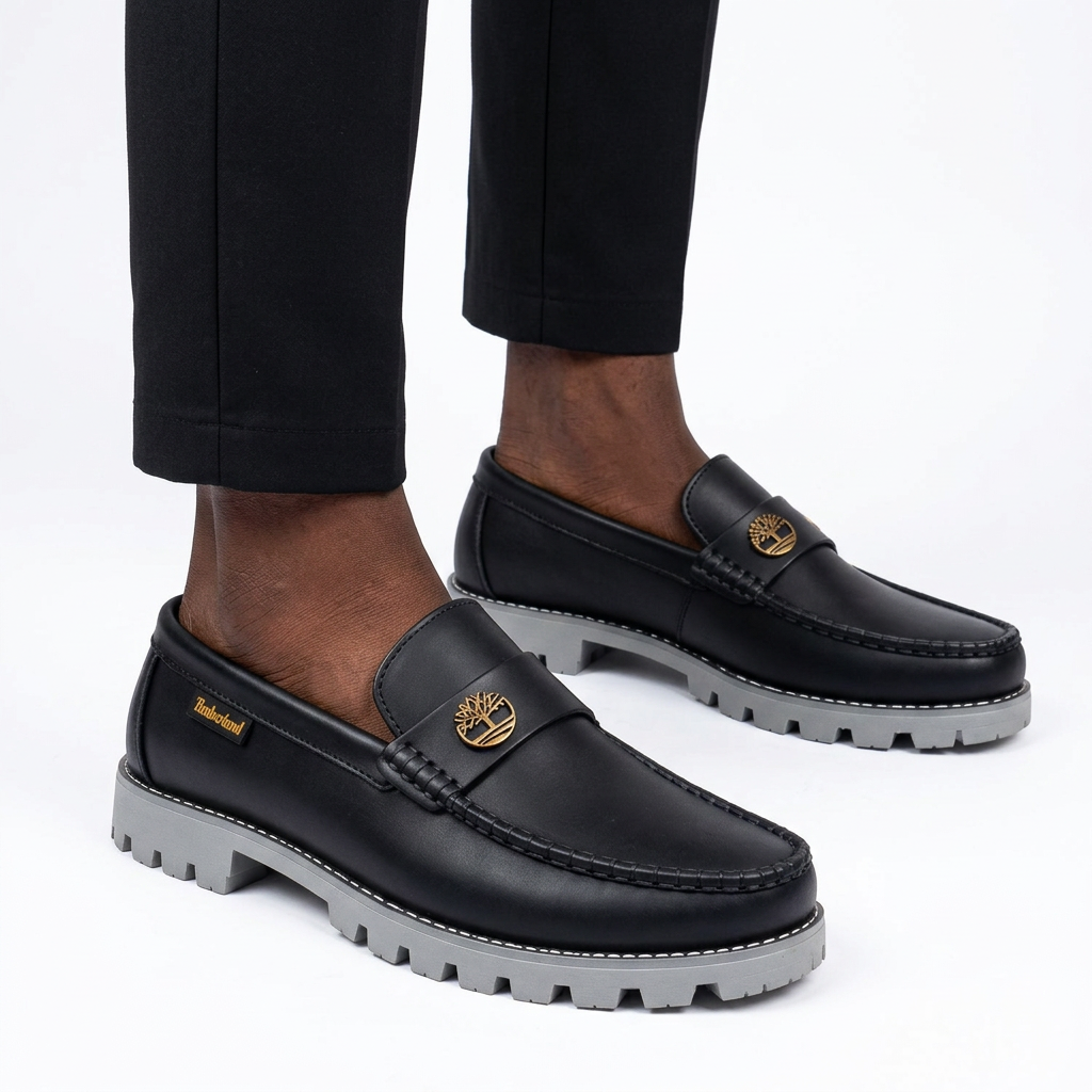 Male model wearing black gold-emblem grey lug-sole loafers, both shoes visible on white