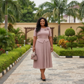 Nigerian female model wearing blush mauve monochrome pleated midi gown with flower corsage in upscale garden setting