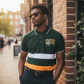 STEEZU Varsity Hunter Green Polo with orange and white chest stripes. Short sleeves, collared, and made of cotton. Features a classic collegiate style.